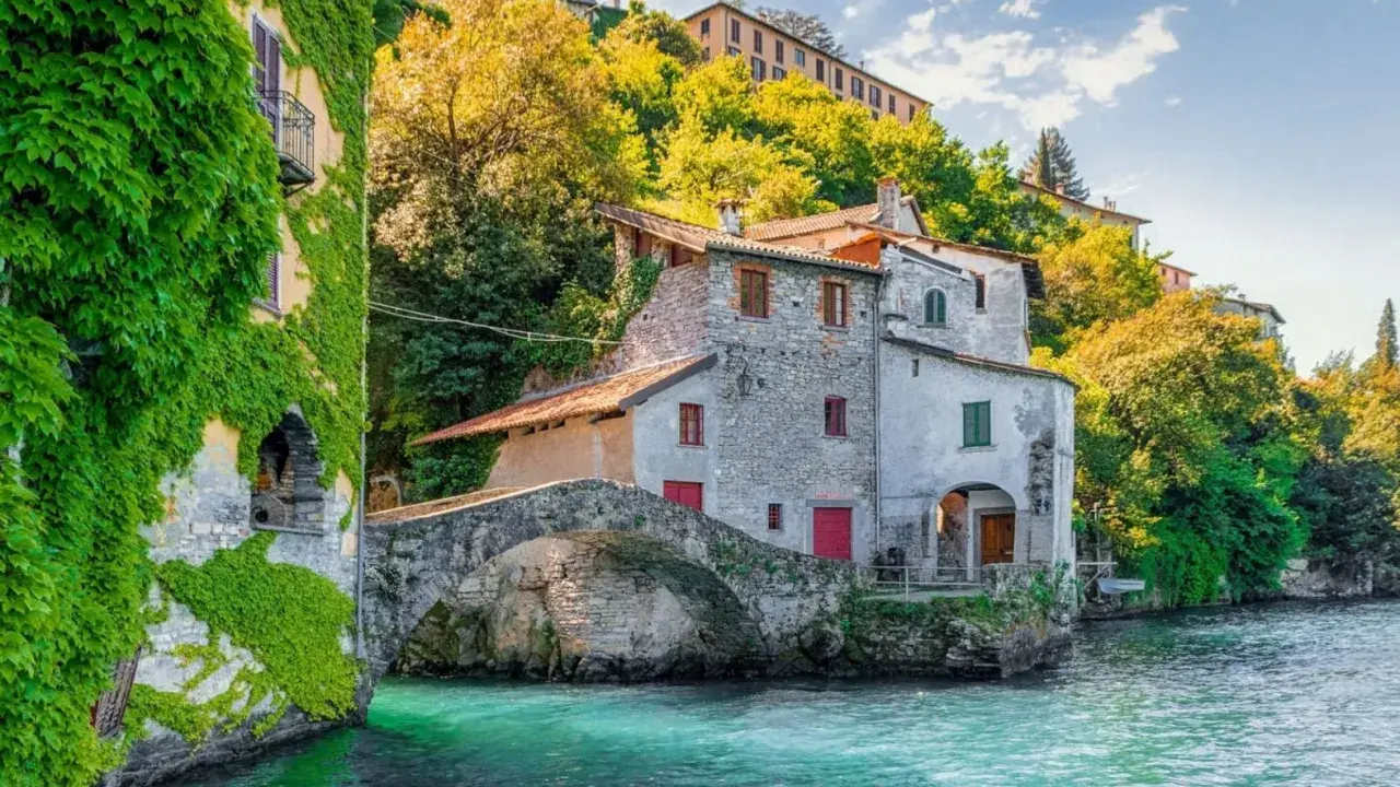 Antica casa in pietra con edera su un lago turchese, un ponte ad arco e colline verdi sullo sfondo, un angolo da fiaba dei castelli Lombardia.