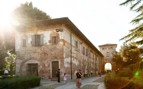 Un sole caldo illumina l'antica **abbazia di Chiaravalle**, con i suoi mattoni a vista e le persiane chiuse. Persone passeggiano nel cortile acciottolato.