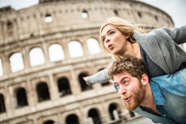 Coppia in una vacanza romantica a Roma, con il Colosseo sullo sfondo. Lui porta lei sulle spalle, entrambi con espressioni sorprese.